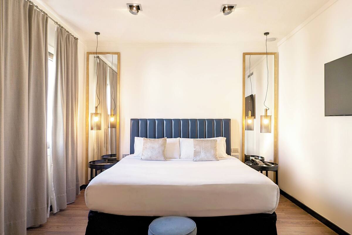 Une chambre d’hôtel avec un grand lit, une table de chevet, un miroir et une fenêtre avec des rideaux.