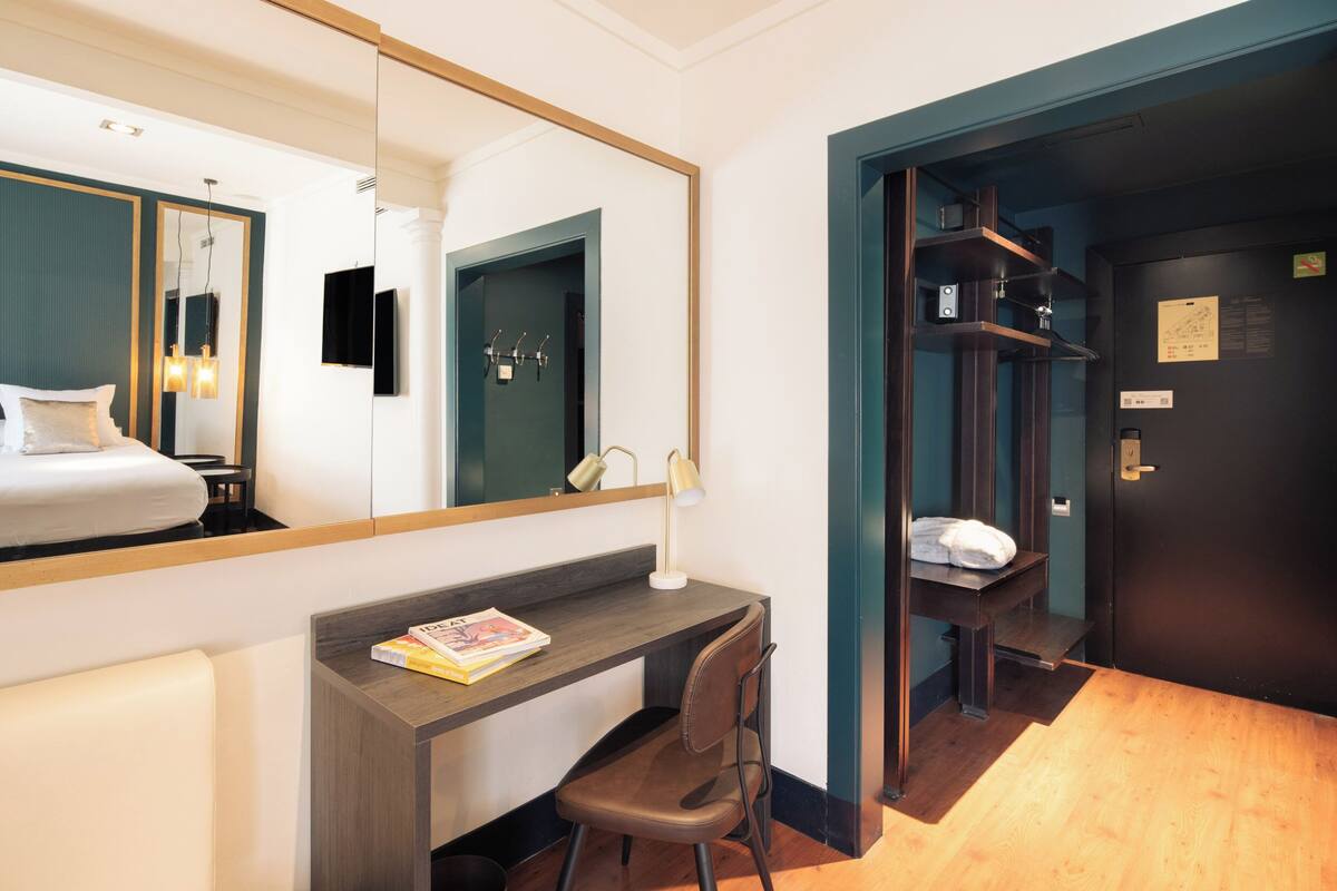 Une chambre d’hôtel comprenant un lit, un bureau avec une chaise, un miroir, une lampe et une porte donnant sur une salle de bain.