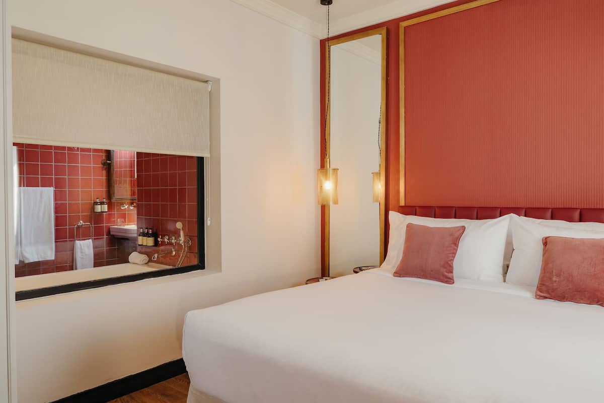 Une chambre d’hôtel avec un lit, un grand miroir, une fenêtre donnant sur la salle de bain et un mur d’accent de couleur rouge.