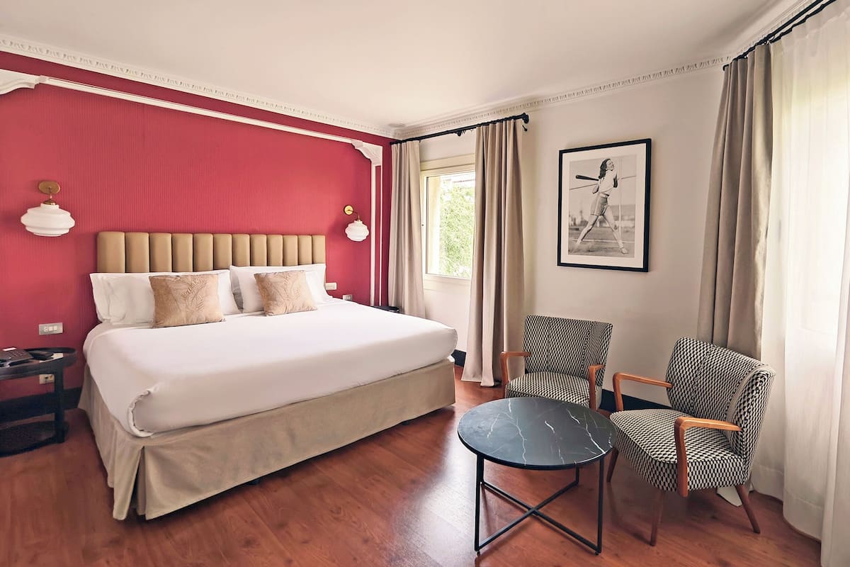 Une chambre d’hôtel avec un grand lit, une petite table, deux chaises, une fenêtre avec des rideaux et un tableau encadré au mur.