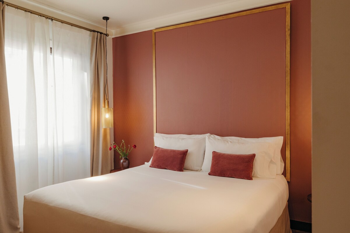 Habitación de hotel con una cama grande, una mesita de noche, un jarrón con flores y una ventana con cortinas.