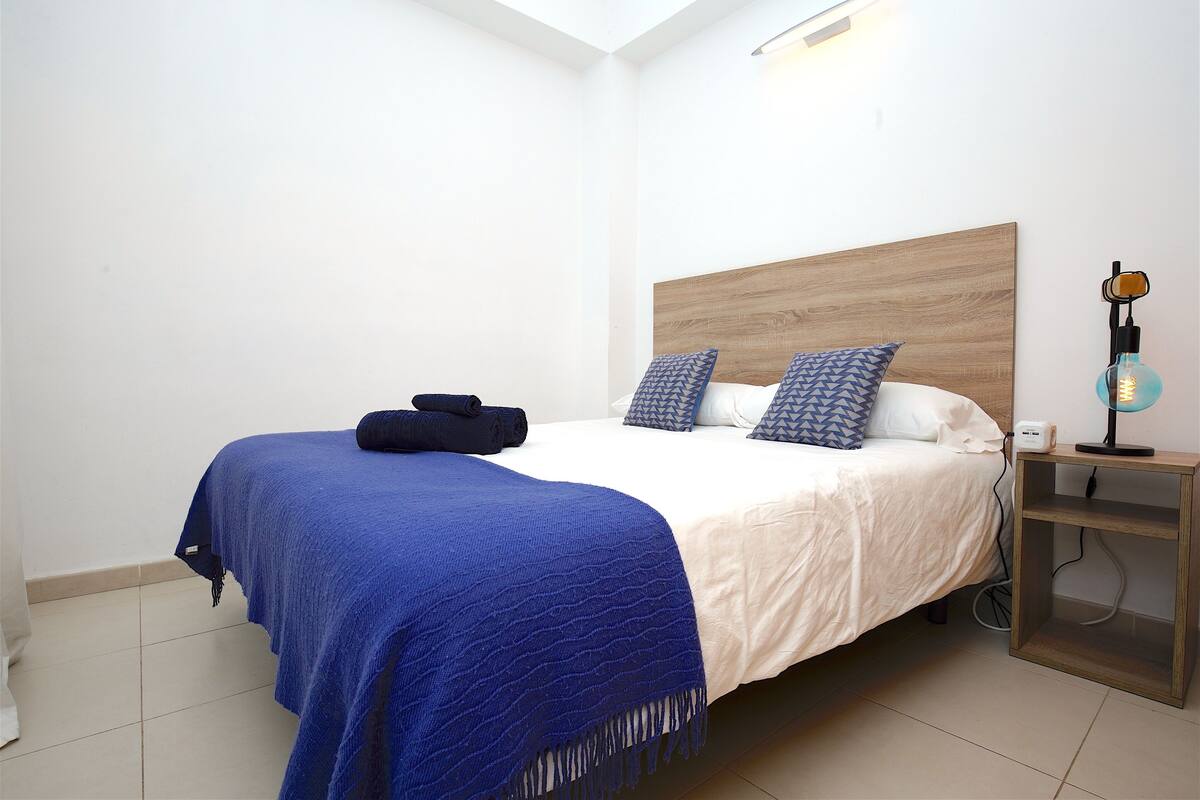 A neatly made bed with a blue blanket and pillows, a bedside table with a lamp, and a wooden headboard.