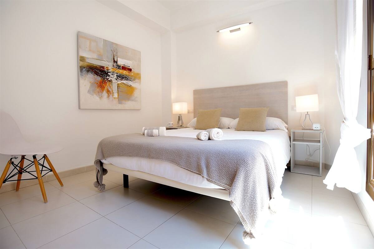 A modern hotel room with a large bed, two bedside tables, a wall-mounted abstract painting, and a view of the outdoors.