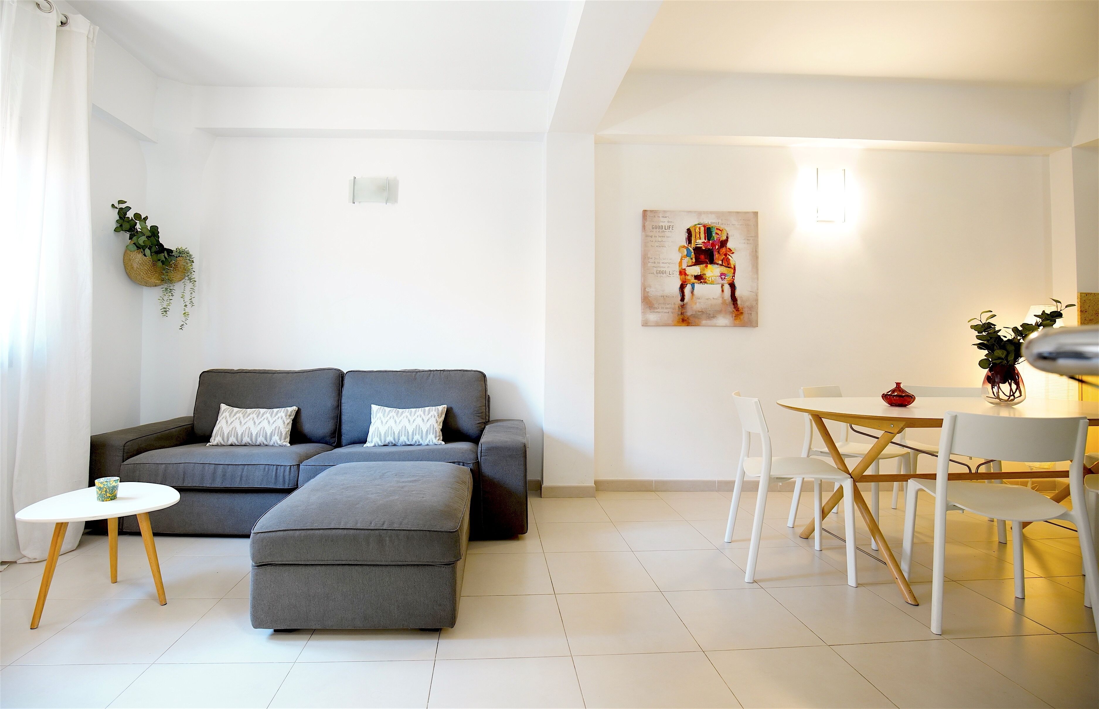 A modern living room with a grey sofa, a small round table, and a dining area with white chairs and a wooden table.