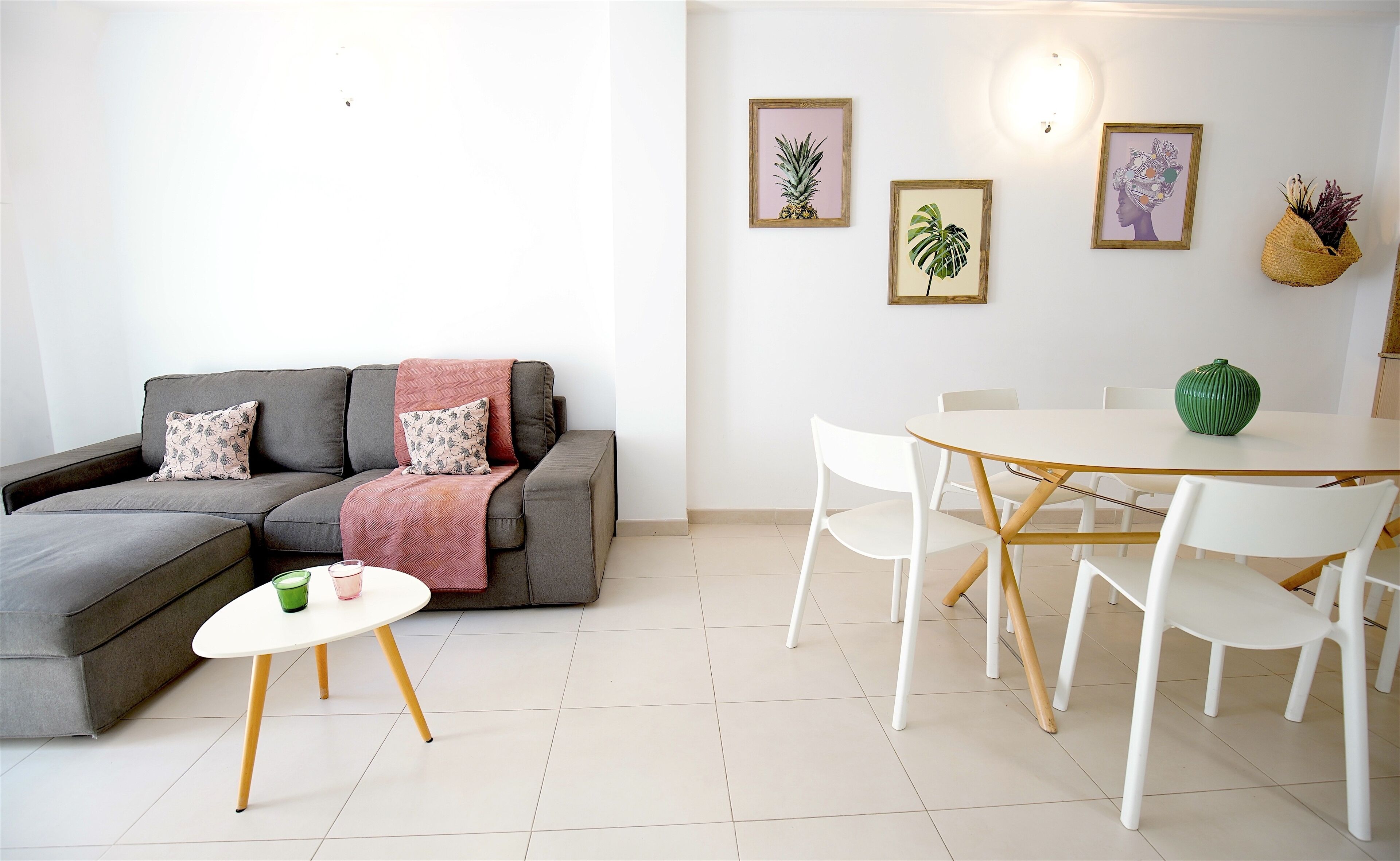 Un salon moderne avec un canapé d’angle gris, une petite table ronde avec deux verres, et un coin repas avec une table et des chaises blanches.