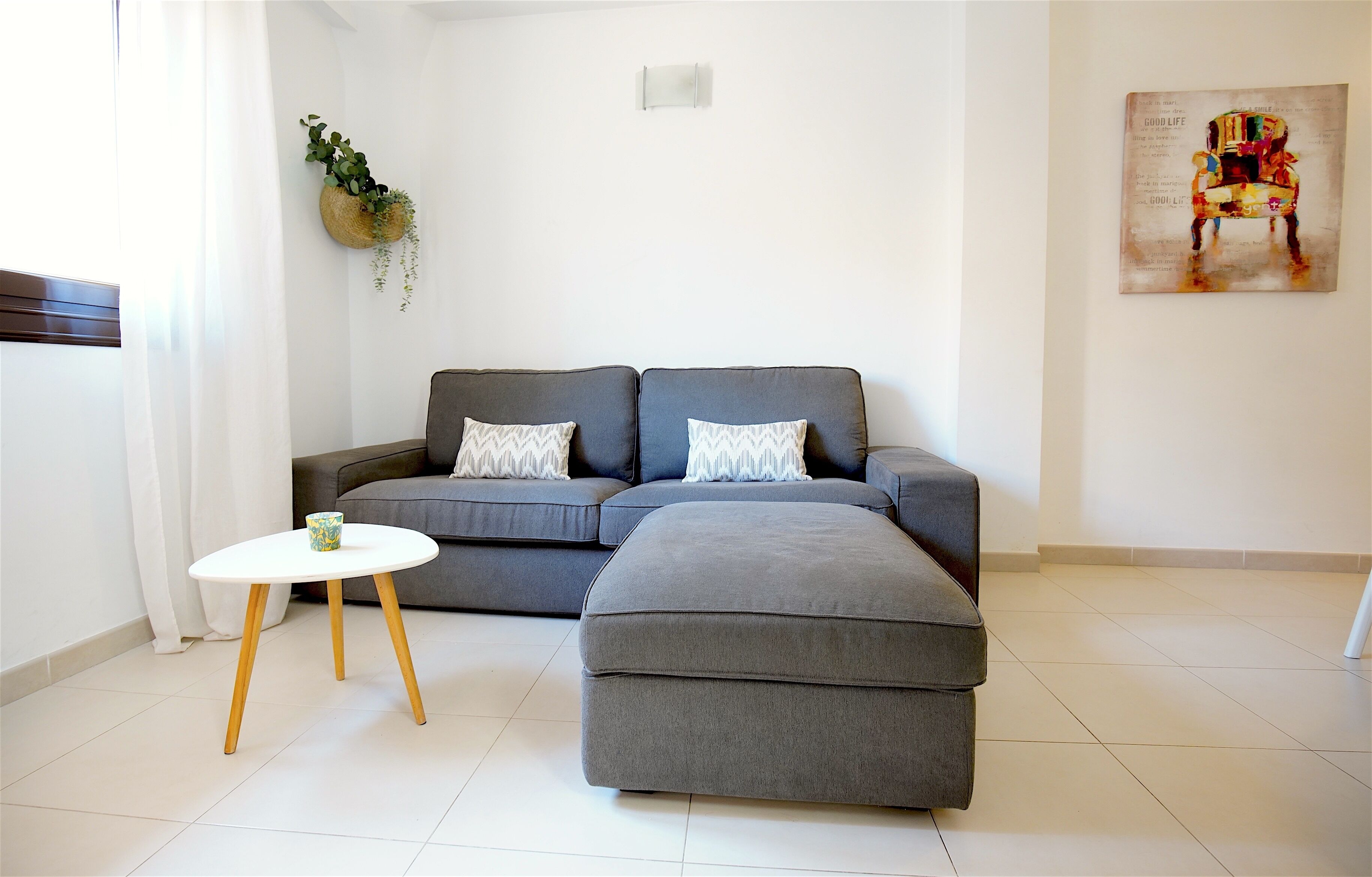 A modern living room with a grey sectional sofa, a small white table with wooden legs, and a decorative wall hanging.