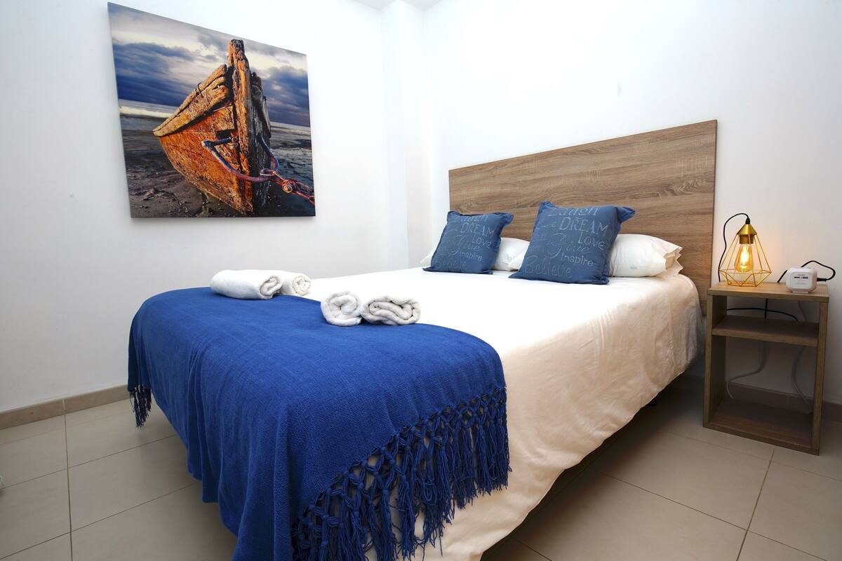 A neatly made bed with a blue blanket and pillows, a wooden headboard, a wall-mounted painting of a boat, and a bedside table with a lamp.