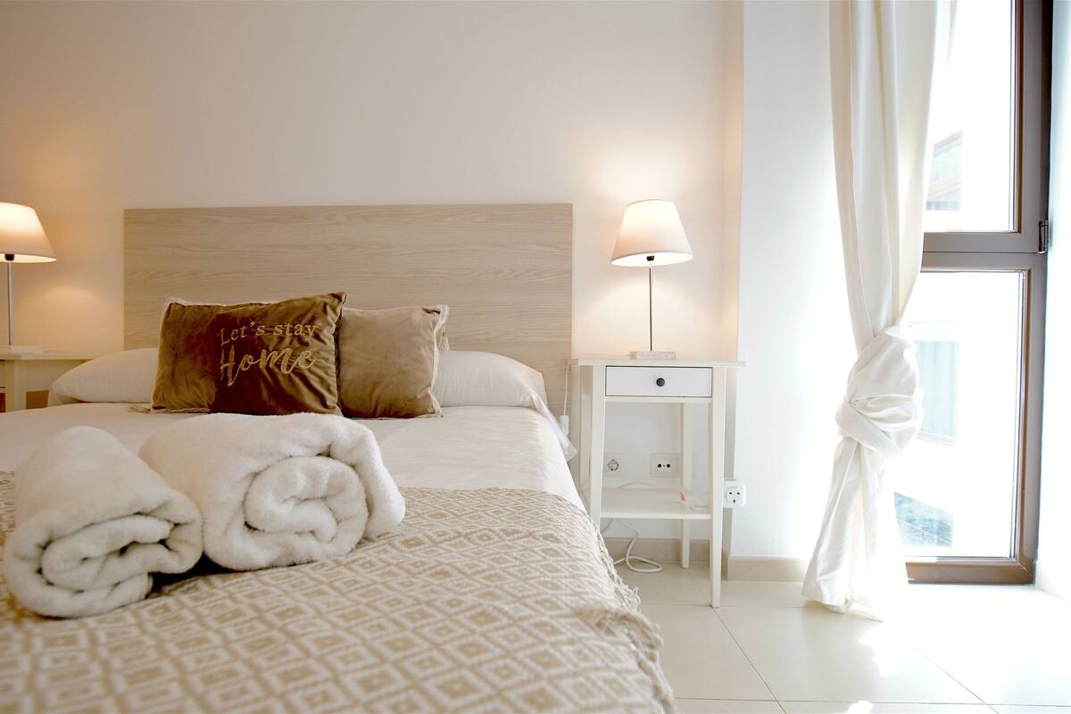 A neatly made bed with rolled towels, a headboard, two bedside tables with lamps, and a window with curtains.