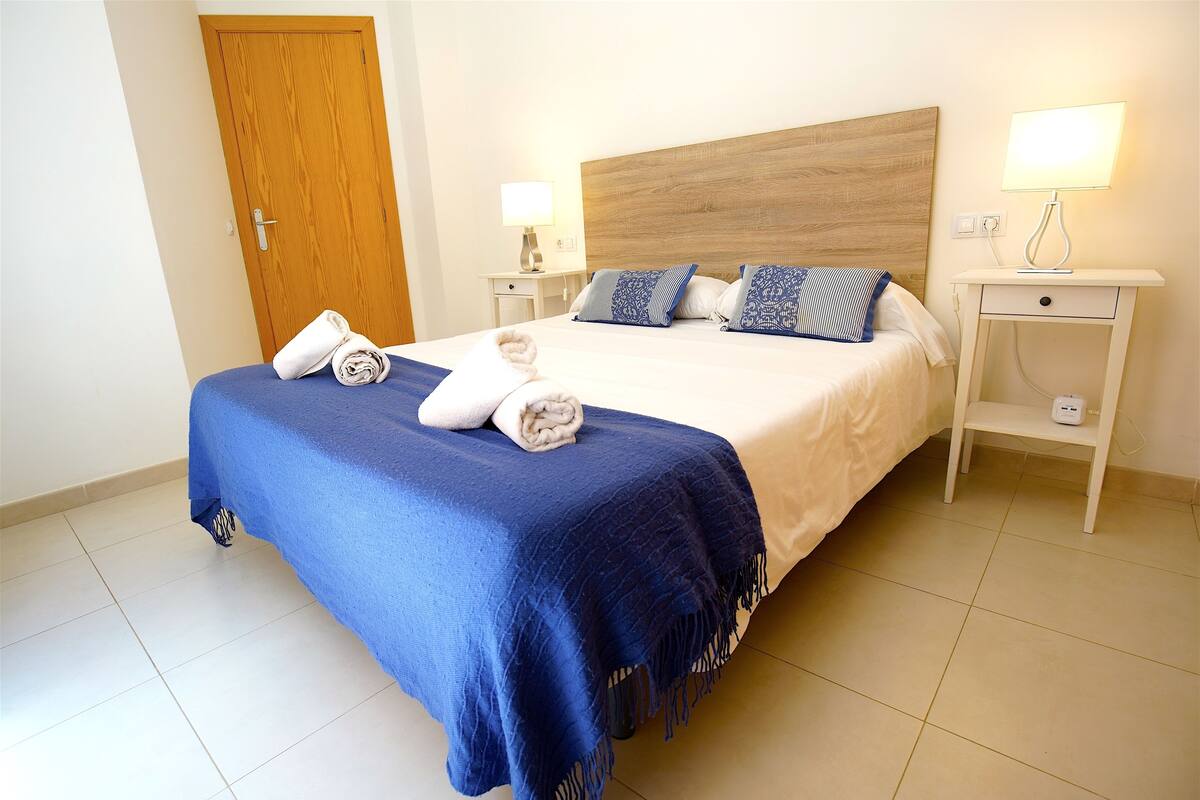 A neatly made bed with a blue bedspread and two pillows, a wooden door, and two bedside tables with lamps.