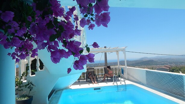 Un espace piscine offrant une vue sur le paysage, une pergola ornée de fleurs violettes et une structure de bâtiment blanche.