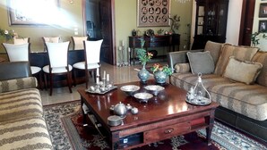 Un salon bien aménagé, avec une table basse en bois, des canapés à motifs et un coin repas comprenant des chaises blanches et un lustre.