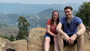 Twee personen zitten op een rots, met uitzicht op een vallei en bergen op de achtergrond.