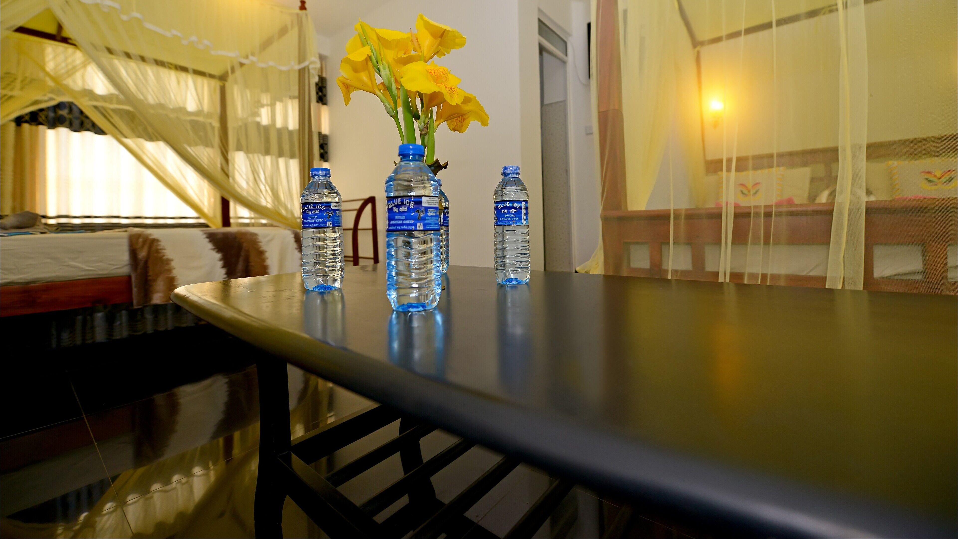 Een kamer met een bed, een tafel met waterflesjes en een vaas met gele bloemen.