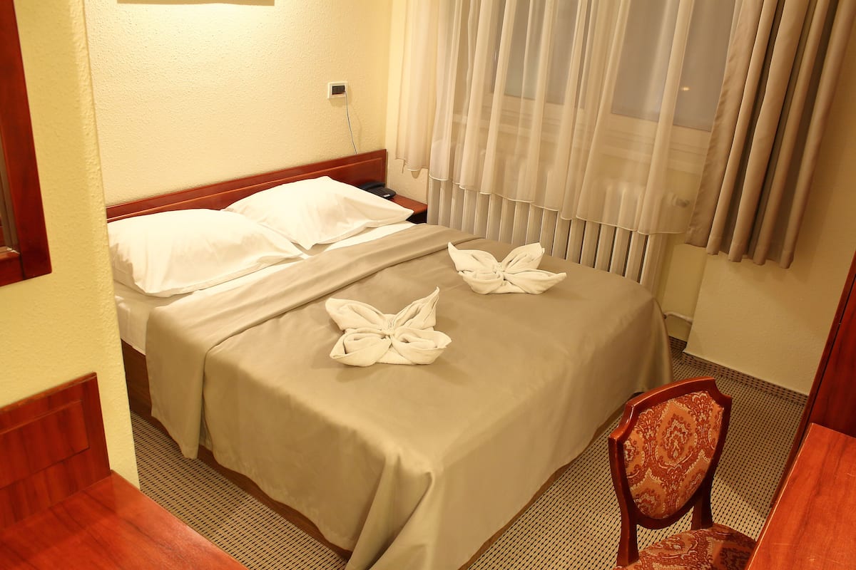 Quarto de hotel com duas camas, cabeceira de madeira, mesa de cabeceira, uma cadeira e uma janela com cortinas leves.