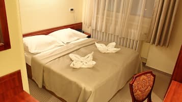 A hotel room with two beds, a wooden headboard, a bedside table, a chair, and a window with sheer curtains.
