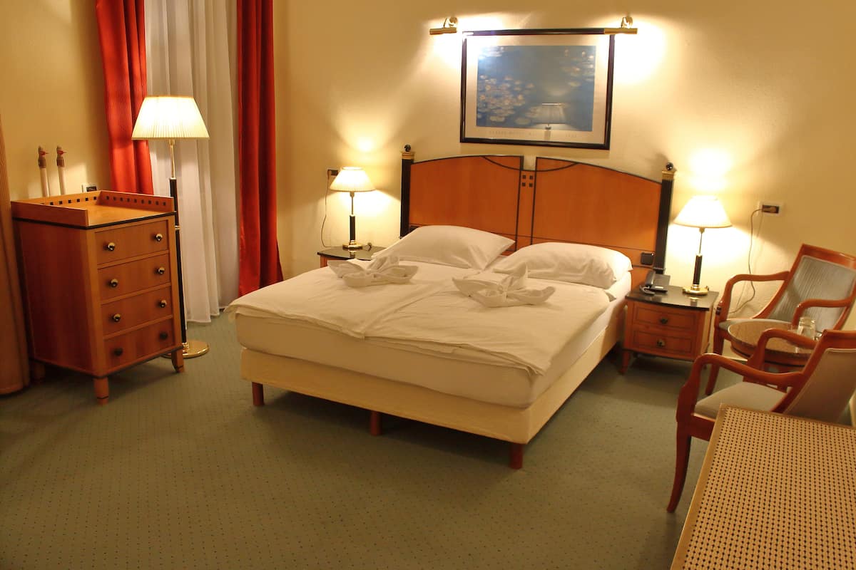 Quarto de hotel com uma cama grande, duas mesas de cabeceira com luminárias, um aparador de madeira, uma cadeira e um quadro na parede.