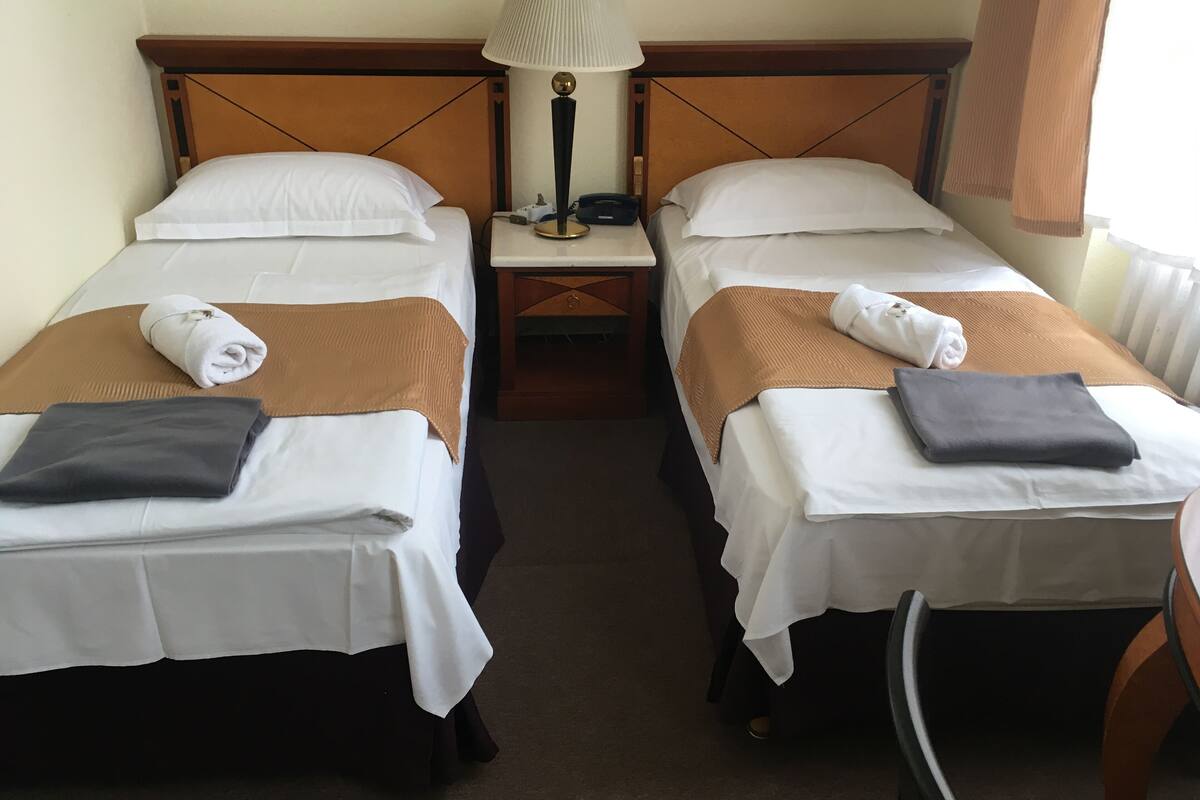 Quarto de hotel com duas camas de solteiro, cada uma com um cobertor marrom e uma toalha enrolada. Há uma mesa de cabeceira com abajur entre as camas.