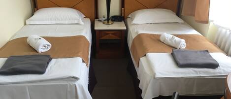 A hotel room with two single beds, each with a brown blanket and a rolled towel. There is a nightstand with a lamp between the beds.