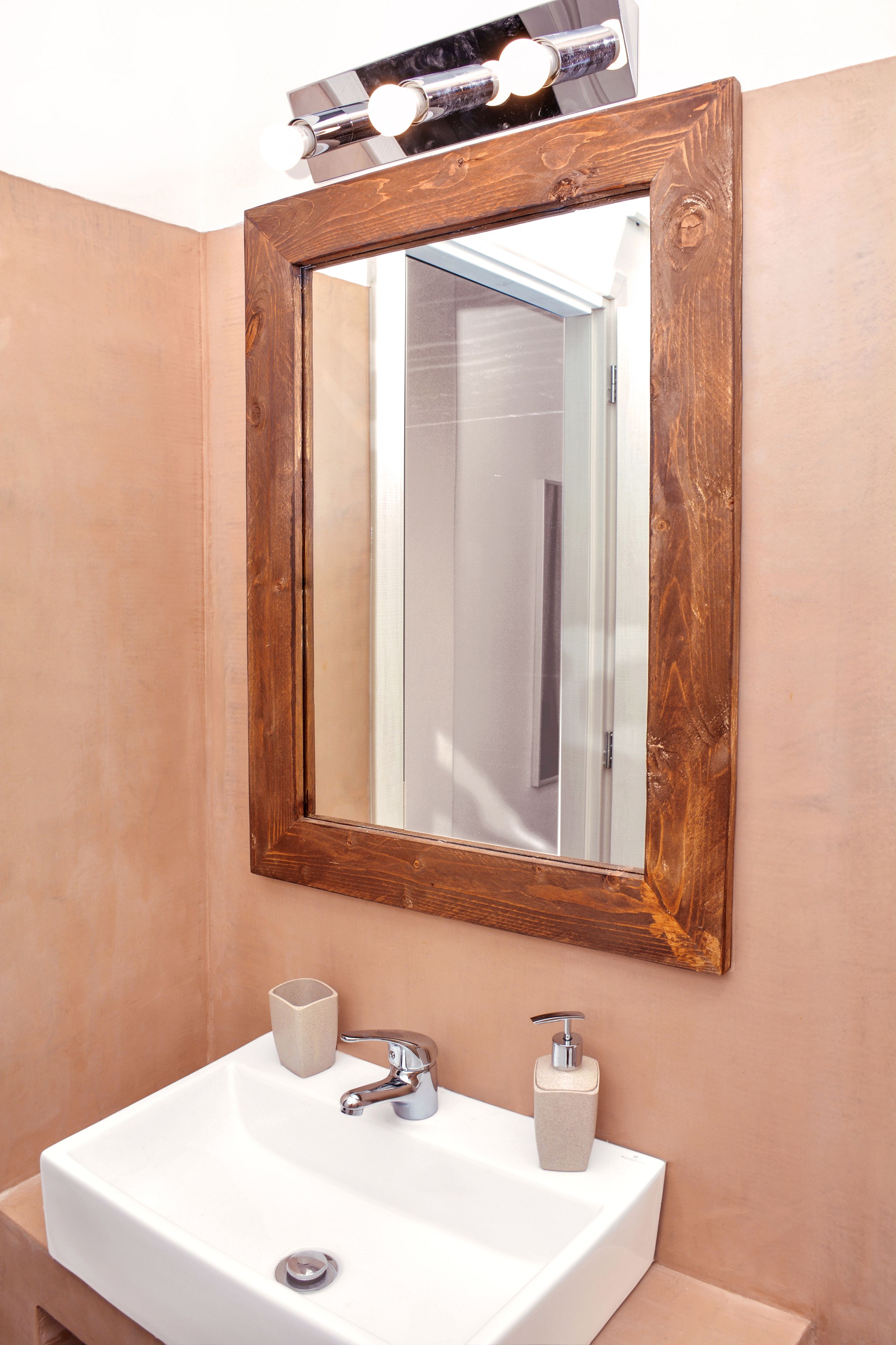 A bathroom with a wooden-framed mirror, a white sink, a soap dispenser, and a cup.