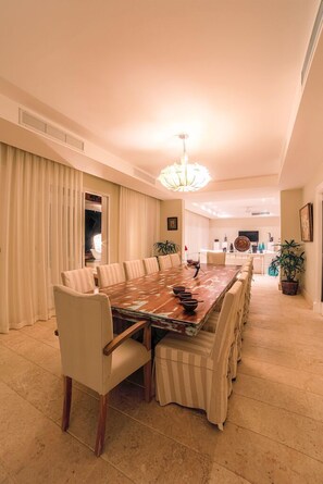 Uma ampla área de jantar com uma longa mesa de madeira, rodeada por cadeiras acolchoadas, um candelabro e plantas em vasos.