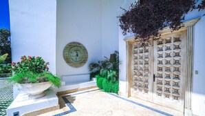 A white building with a tiled entrance, a large decorative door, and a potted plant with red flowers. - Hacienda B-03 (Punta Cana)