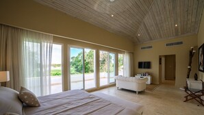 A spacious bedroom with a large bed, a white armchair, and a view of the outdoors through large windows. - Hacienda B-03 (Punta Cana)