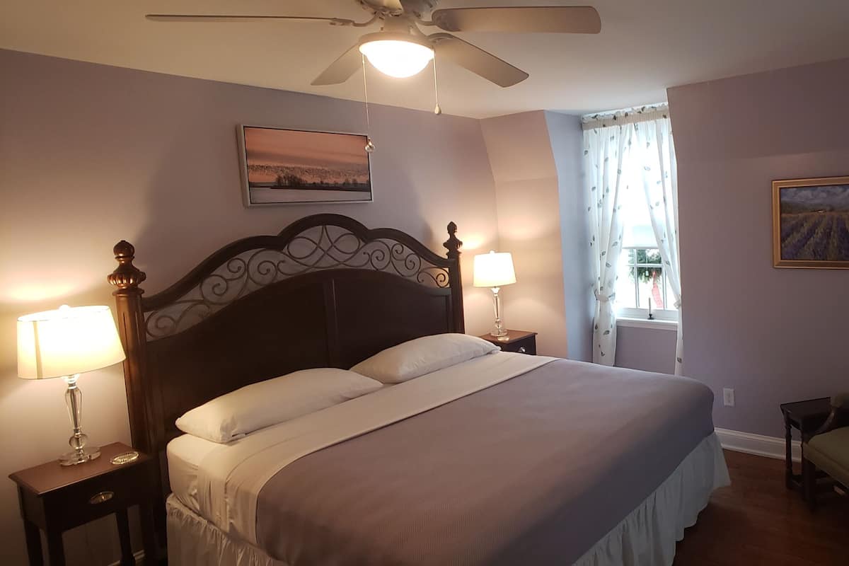 A bedroom with a large bed, two bedside lamps, a ceiling fan, a chair, and two paintings on the walls.