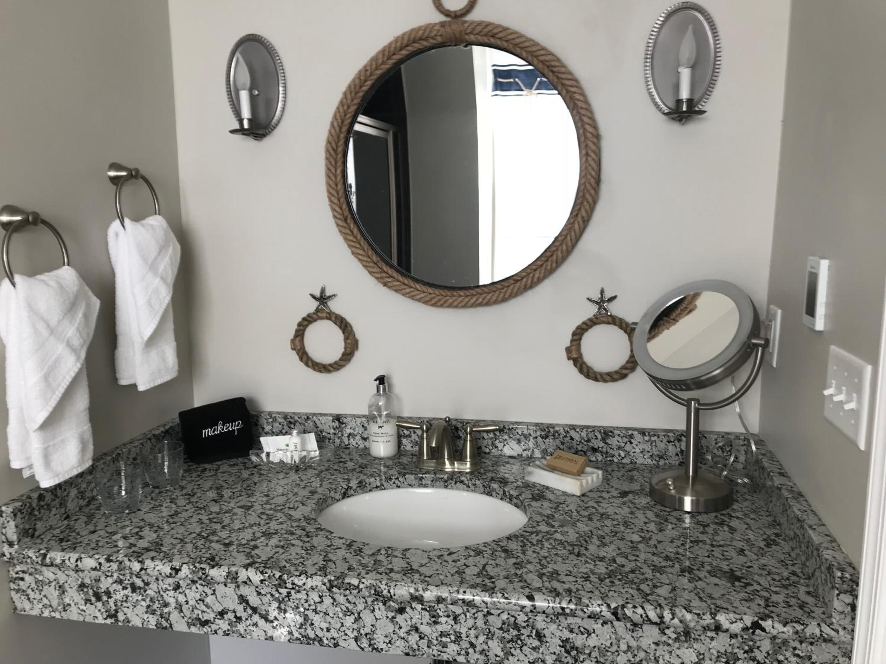 Un baño con encimera de granito, un lavamanos blanco, un espejo redondo con marco de cuerda y dos toalleros, además de un adorno con forma de estrella.