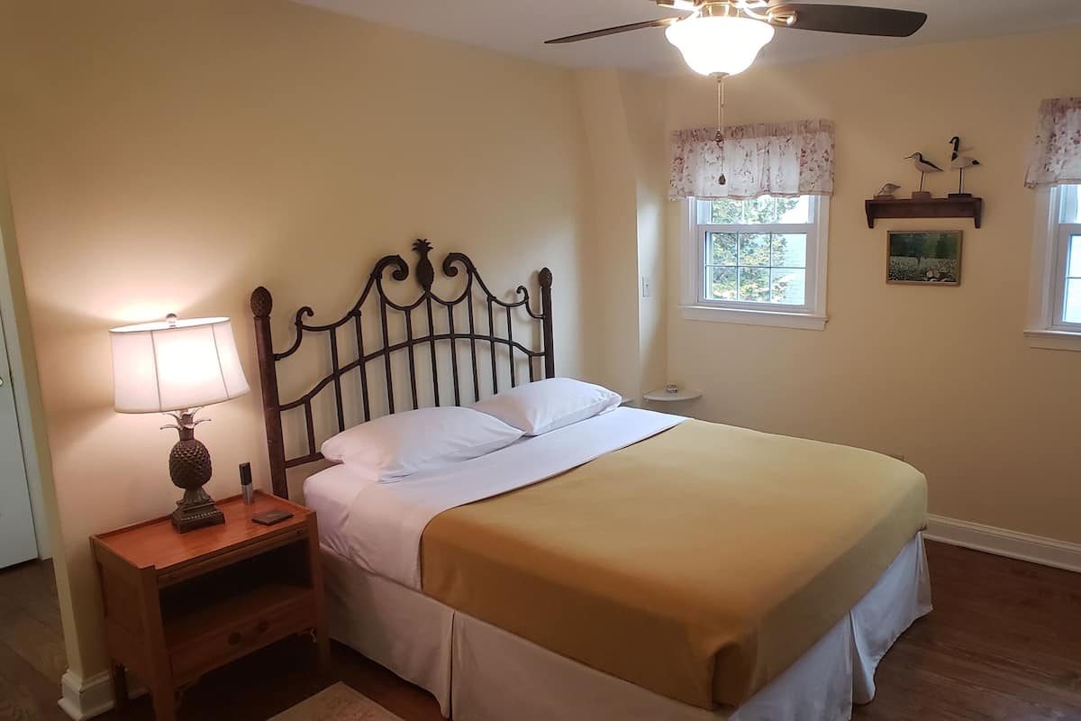 A bedroom with a bed, bedside table, lamp, ceiling fan, windows with curtains, and a dresser.