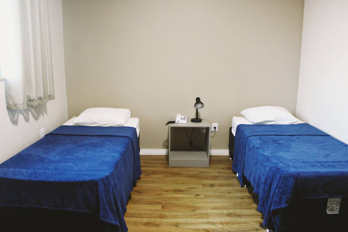 Ein Zimmer mit zwei Einzelbetten, jeweils mit blauer Bettdecke und weißen Kissen, einem kleinen Nachttisch mit Lampe und einer Steckdose an der Wand.