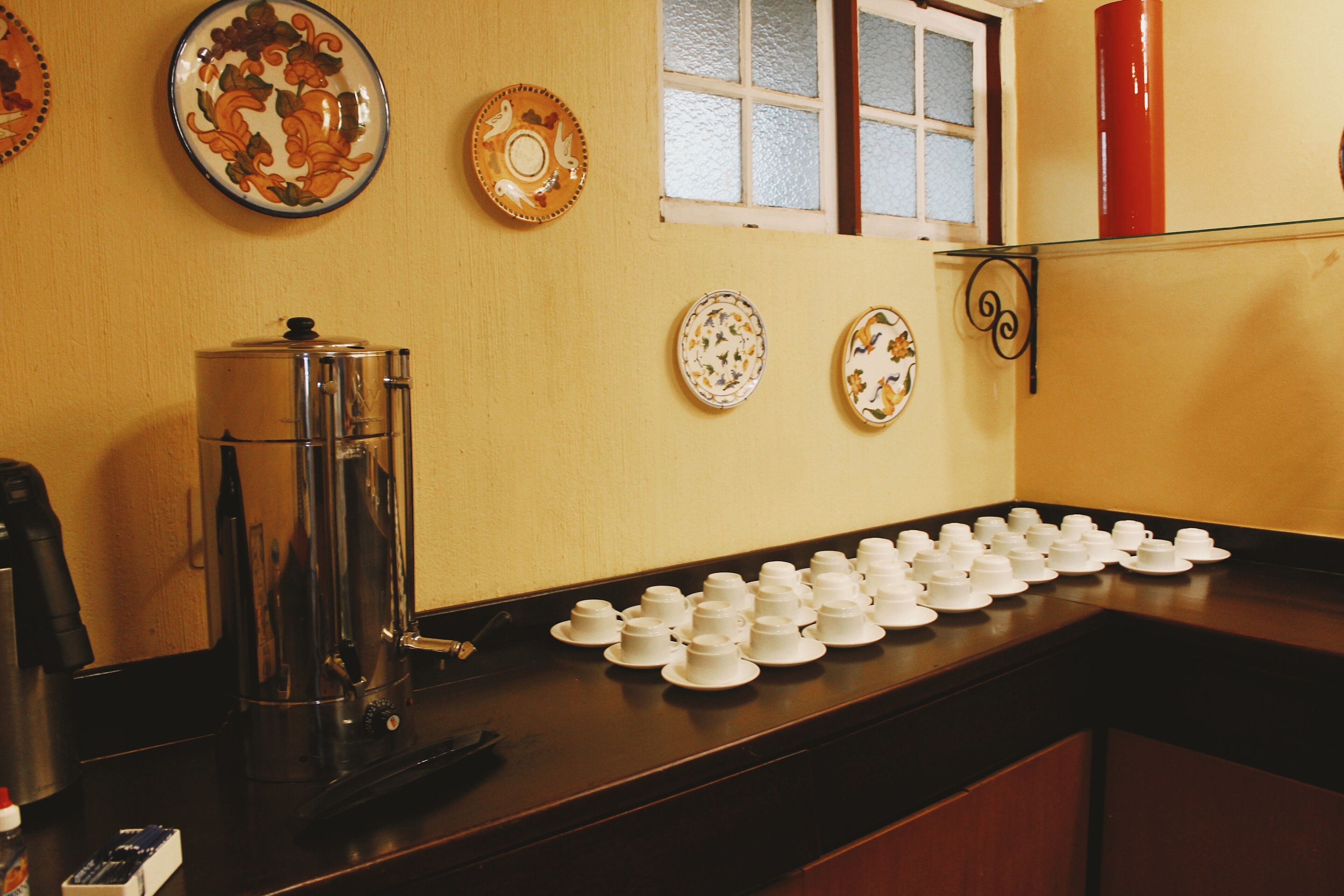 Un comptoir de cuisine avec une machine à café, une rangée de tasses à thé blanches et des assiettes décoratives accrochées au mur.