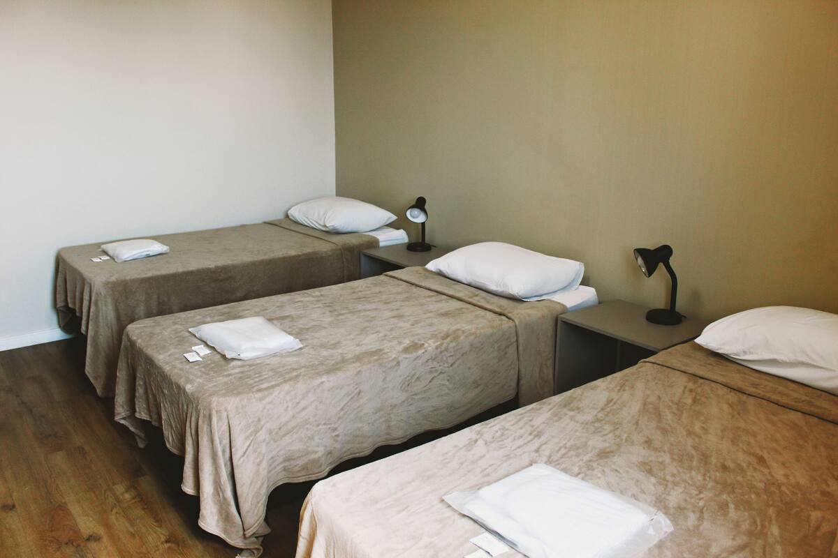 Ein Hotelzimmer mit drei Einzelbetten, jedes mit einem weißen Kissen und einem gefalteten weißen Handtuch. Es gibt zwei Nachttische mit Lampen.