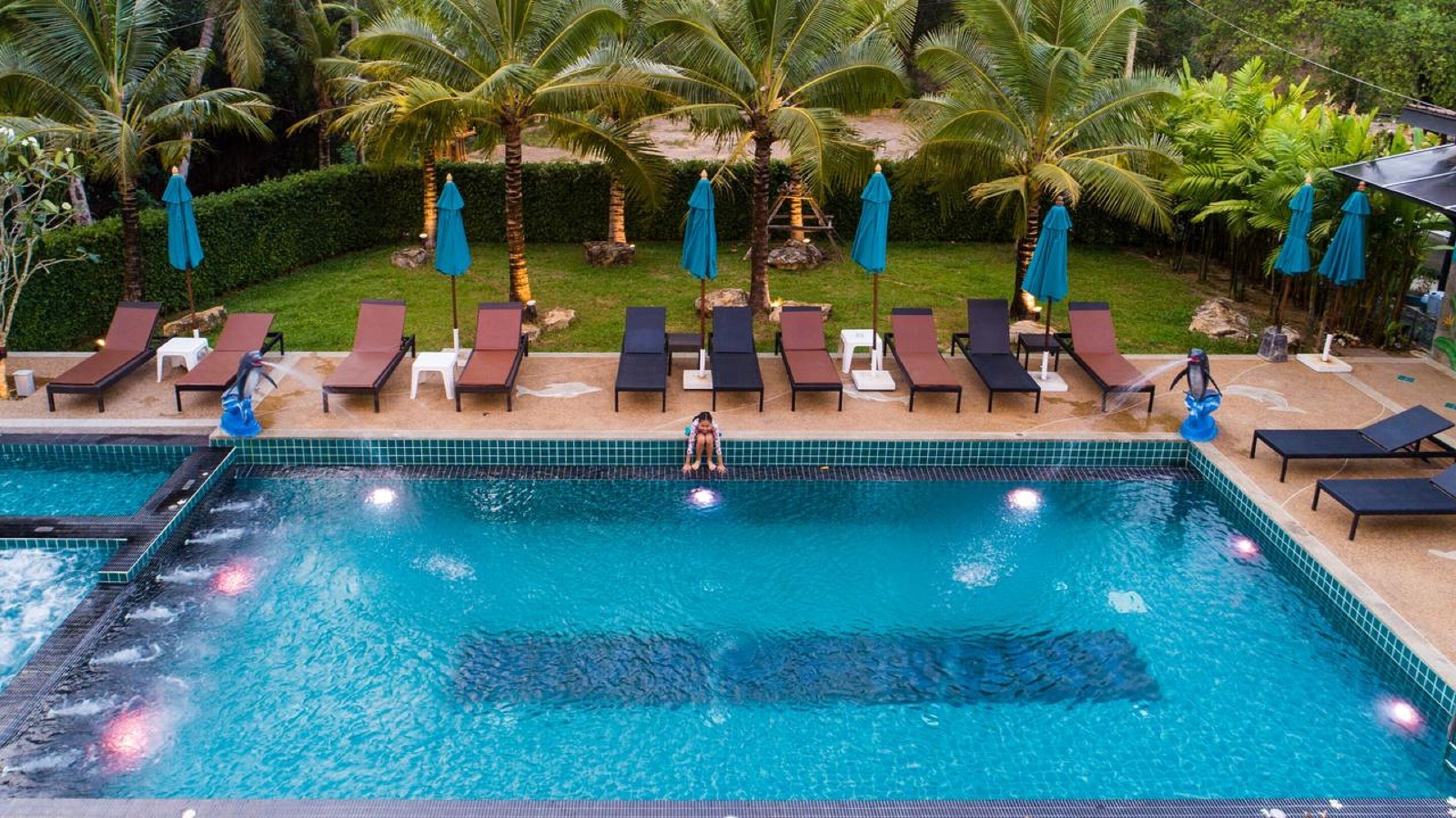 Una piscina con sillas de descanso y sombrillas, rodeada de palmeras y un césped cuidado.