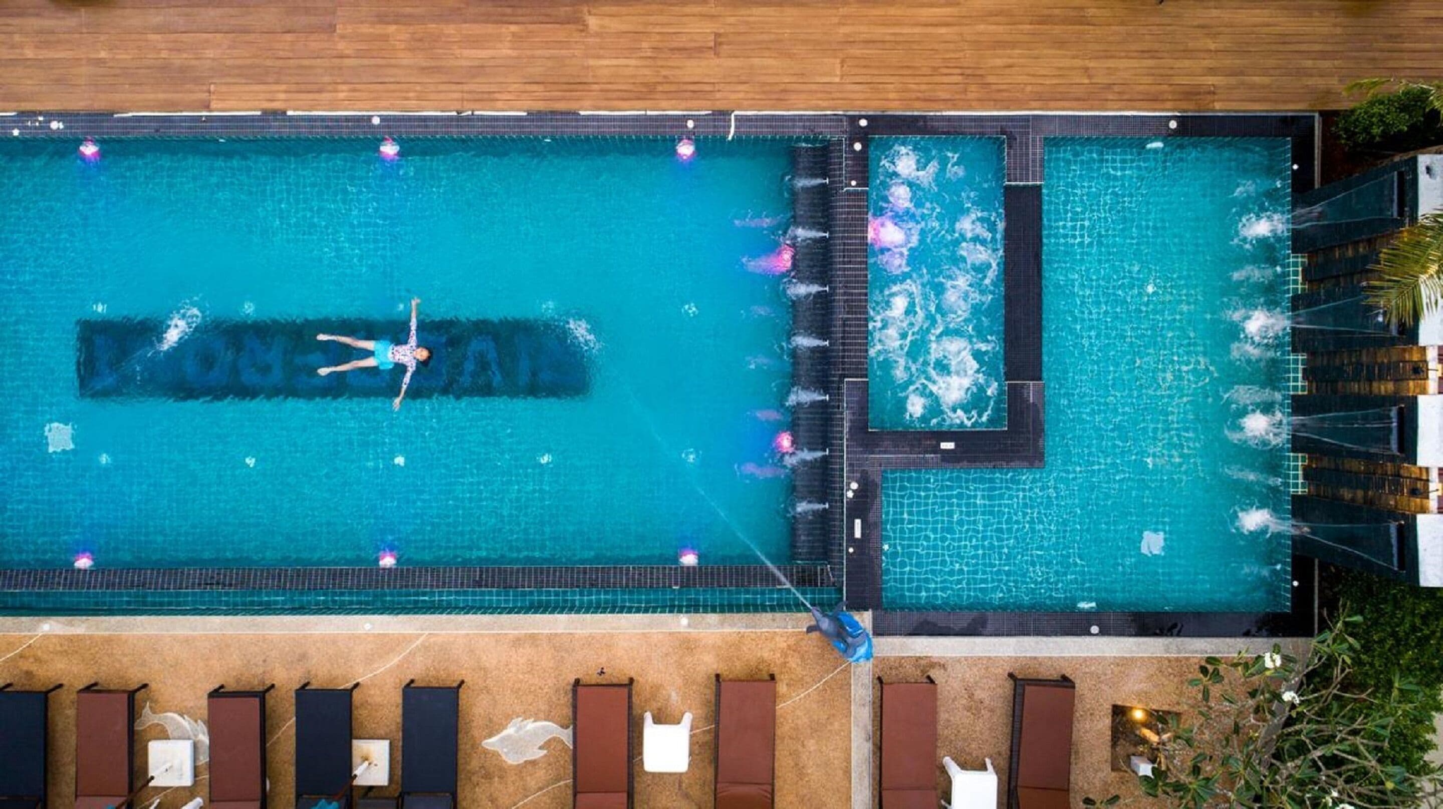 An aerial view of a rectangular swimming pool with a tiled deck surrounding it. There are lounge chairs and a small section of the pool with a different tile pattern.