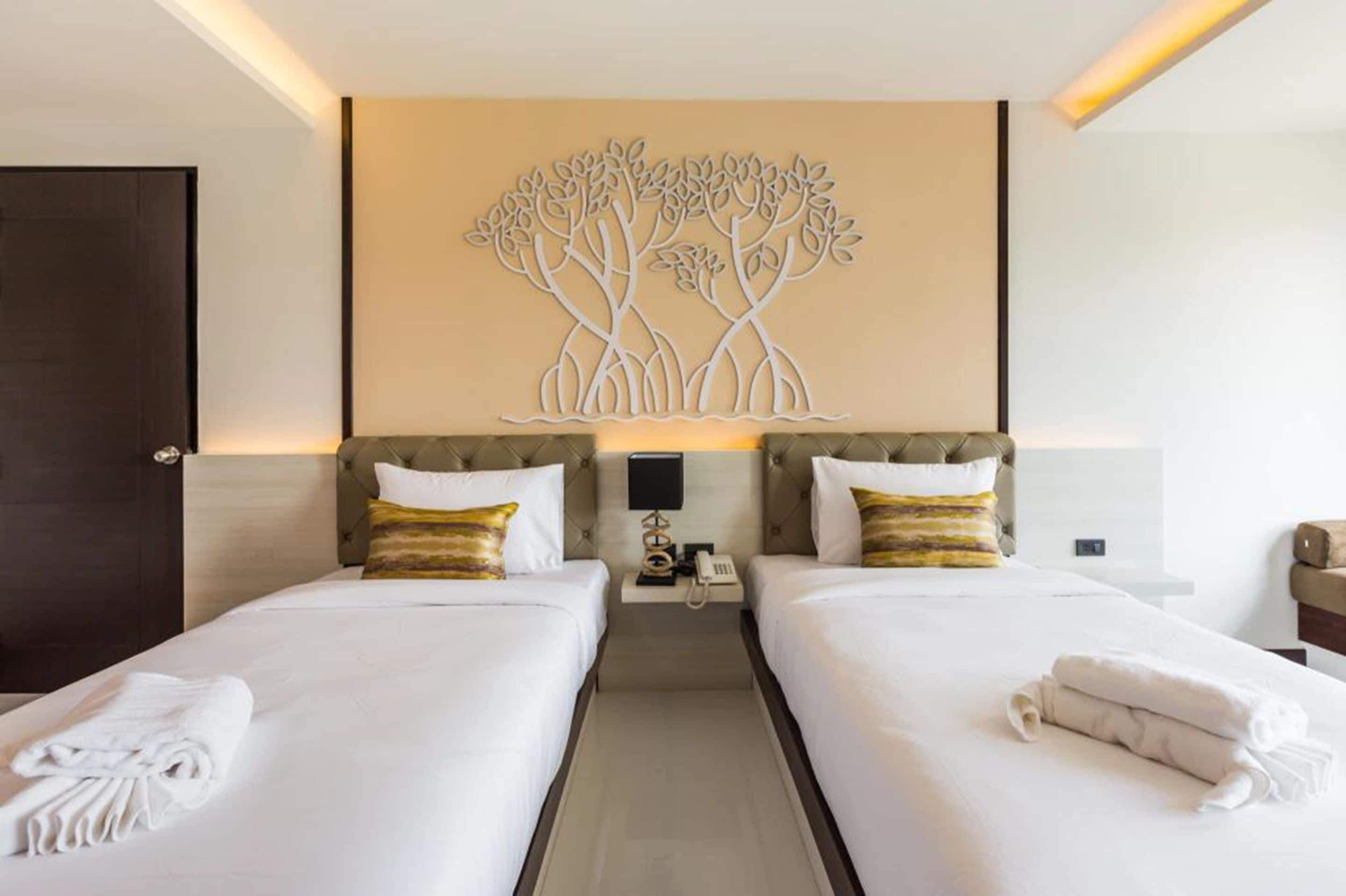 A hotel room with two beds, a wall-mounted artwork of trees, a bedside table with a lamp, and a phone.