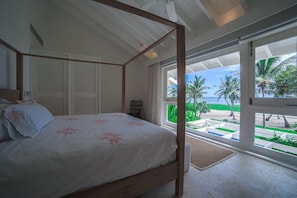 A four-poster bed with a white bedspread and a view of a beach through large windows. - Arrecife 17 (Punta Cana)