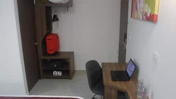 A small room with a bed, a desk with a laptop, a chair, a red suitcase, and a shelf with a pillow and a bag.