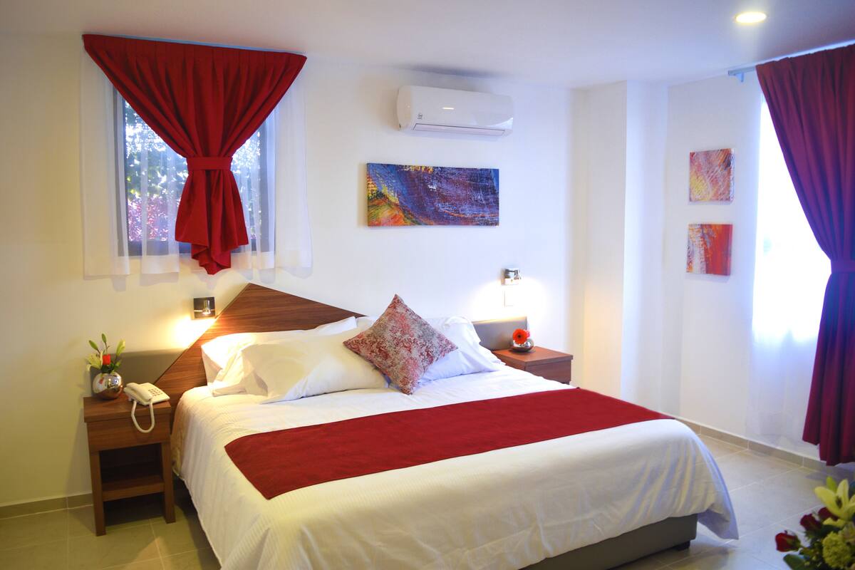 A hotel room with a bed, a nightstand, an air conditioner, and artwork on the wall.