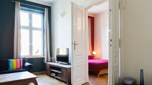 A room with a wooden floor, a television on a stand, a sofa with a colorful pillow, a bed with a red blanket, and a door leading to another room. - Krakowrentals - Stanislas Apartment (Kraków)