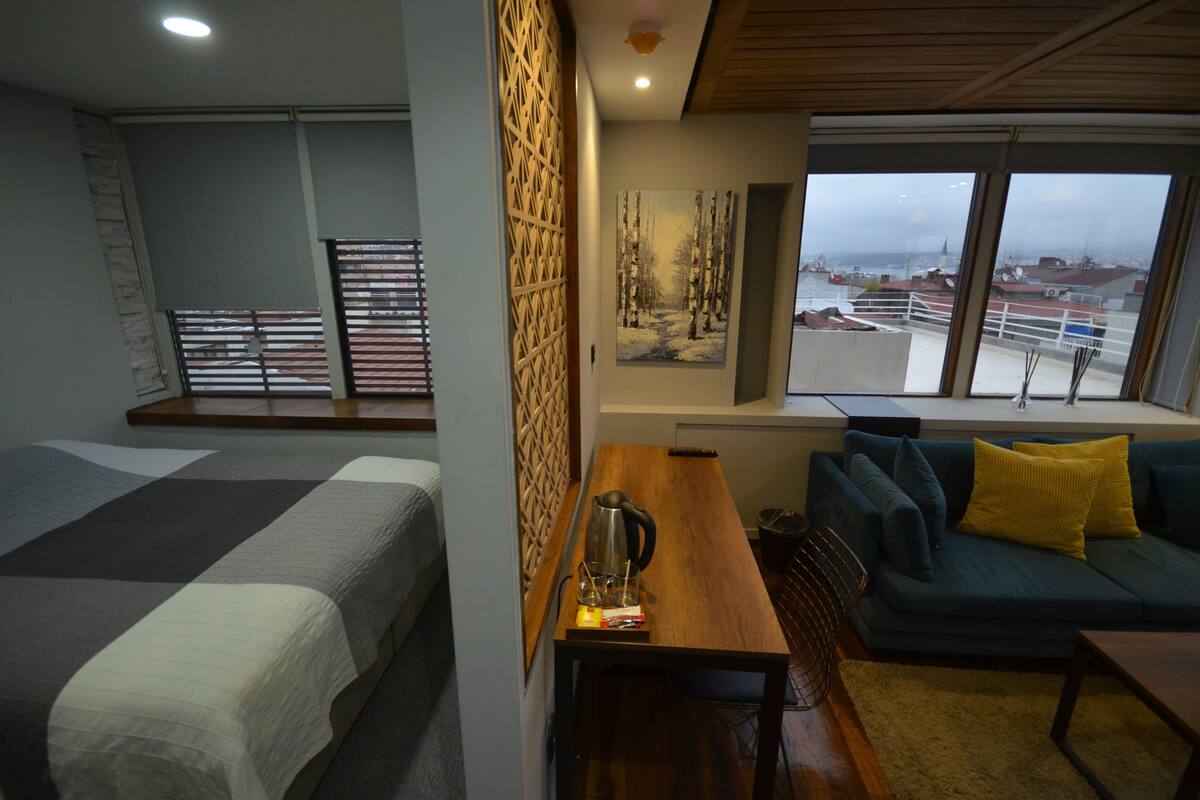 A room with a bed, a wooden table, a sofa, and a painting of a forest.
