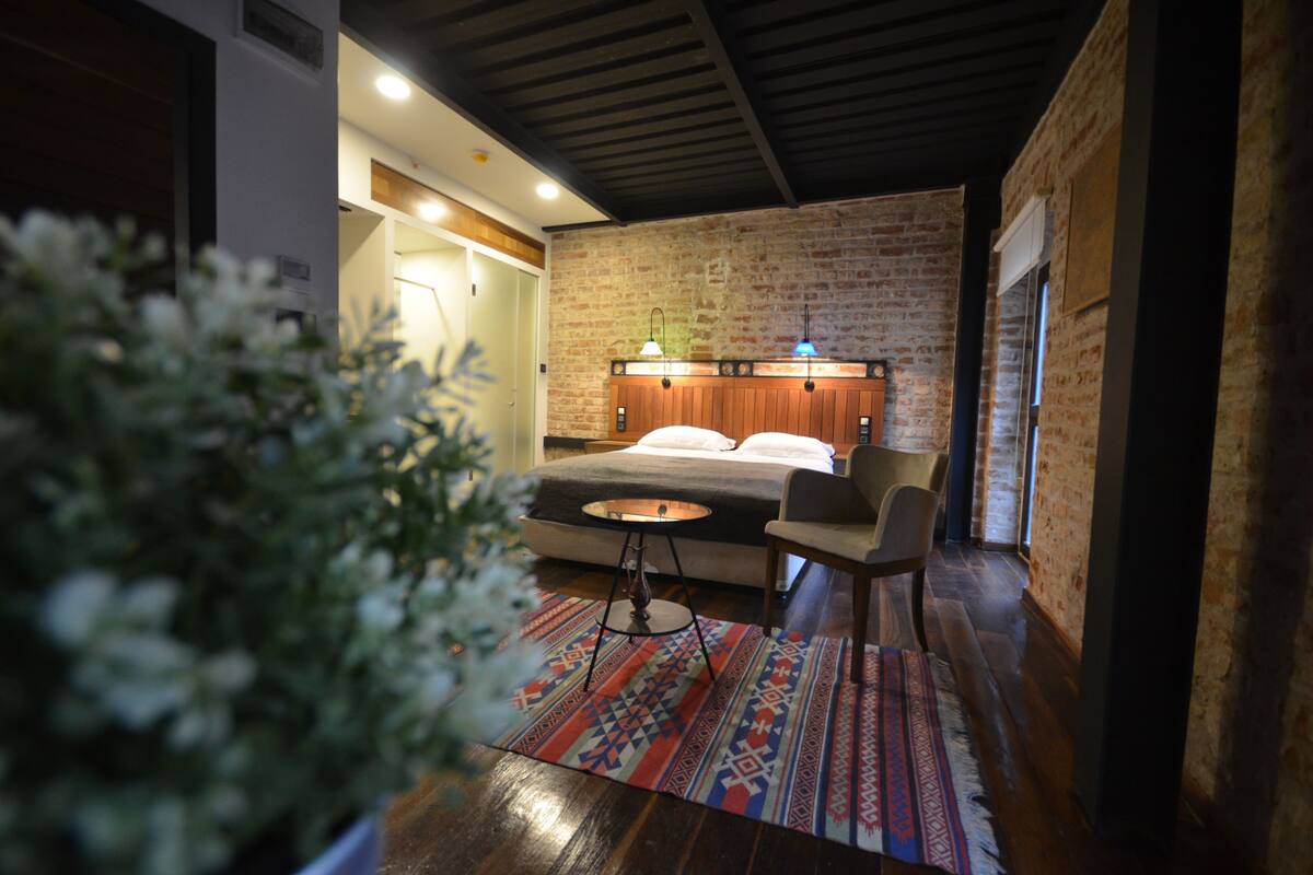 A cozy bedroom with a brick wall, a bed with a wooden headboard, a small round table, a chair, and a colorful rug.