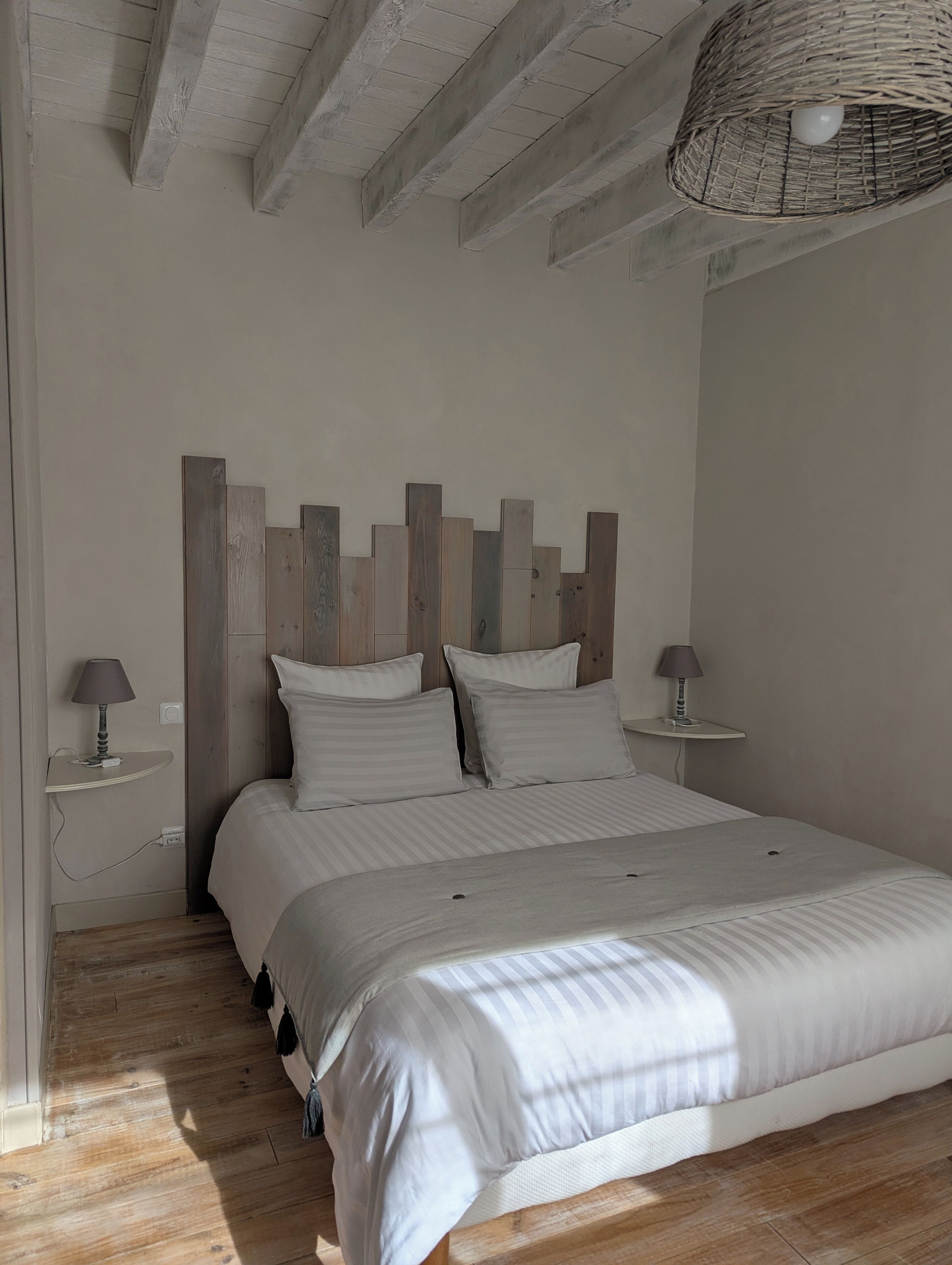 A bedroom with a large bed, two bedside tables with lamps, a woven ceiling light fixture, and wooden beams on the ceiling.