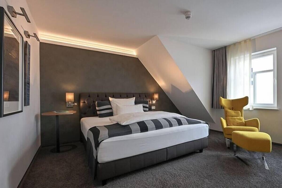 A modern hotel room with a large bed, a small side table, a yellow armchair, and a large window.