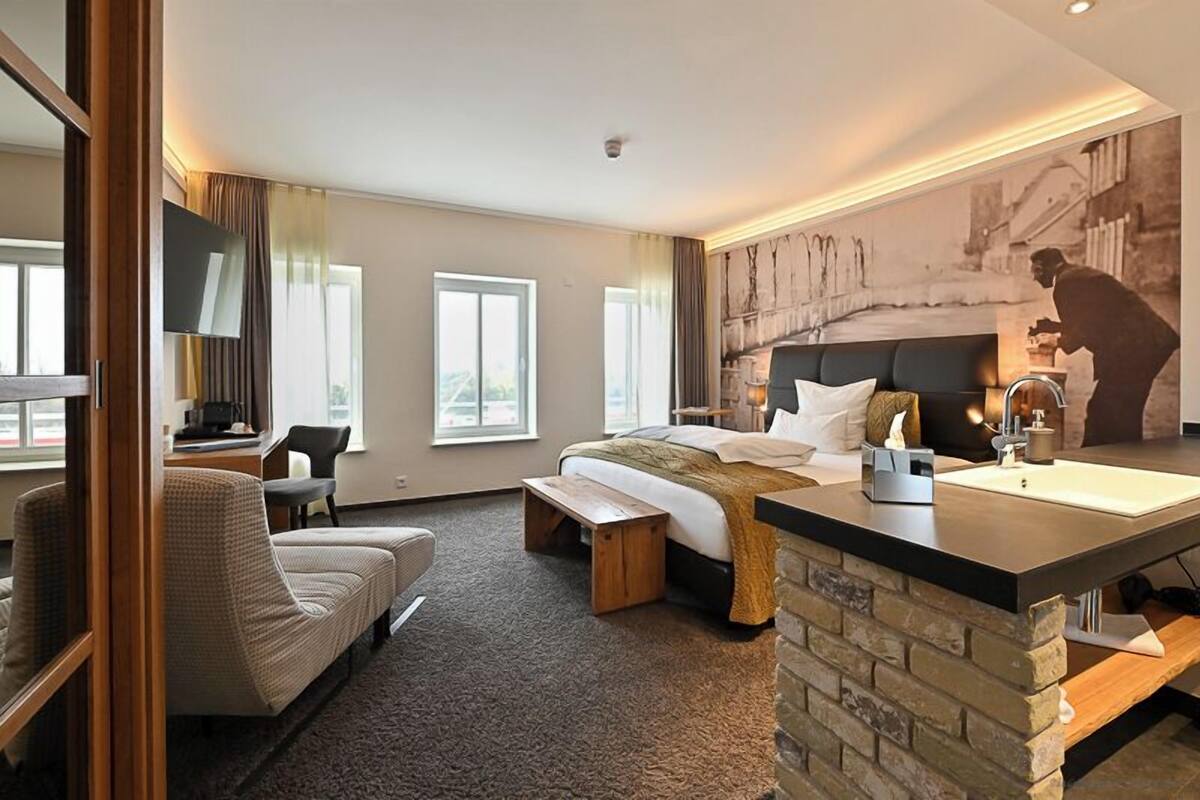 A hotel room with a bed, a desk, a chair, a bench, a small table, and a large window.