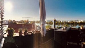 A terrace with outdoor seating, a view of a river, and a cityscape in the background. - Höll am Main (Ruesselsheim)