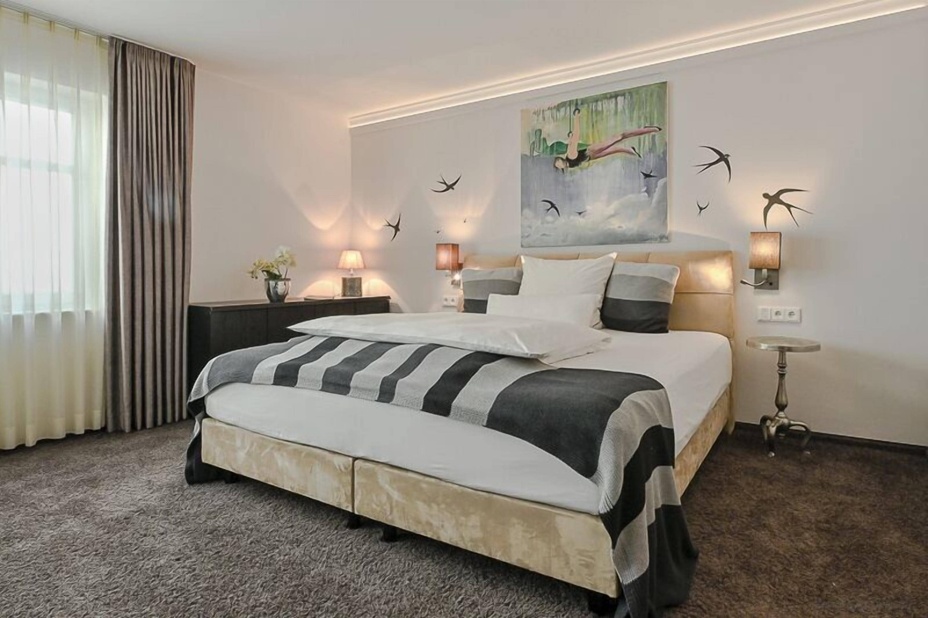 Habitación de hotel con una cama grande, dos mesitas de noche con lámparas, un cuadro en la pared y vistas al exterior.