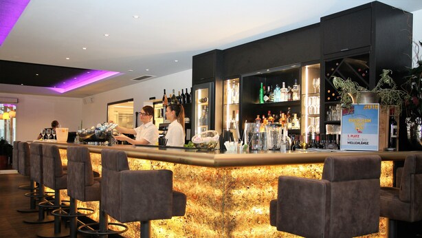 A bar area with a bar counter, stools, and shelves stocked with bottles and glasses. - Höll am Main (Ruesselsheim)