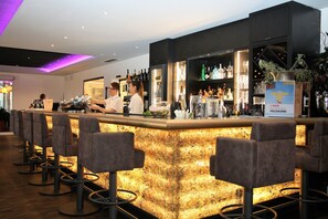 Una zona de bar con mostrador, taburetes y estanterías llenas de botellas y vasos.