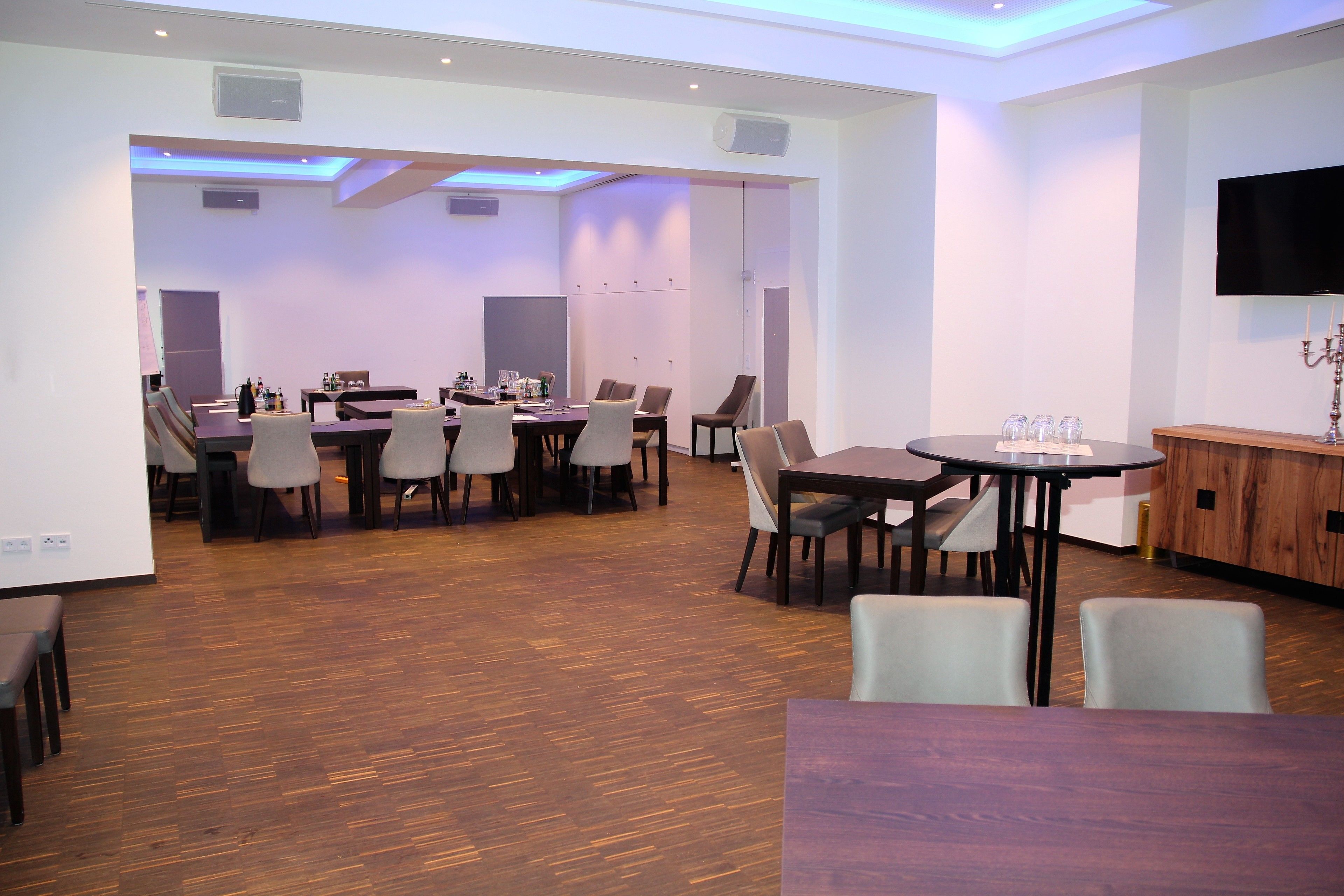 A modern dining area with multiple tables and chairs, a flat-screen TV mounted on the wall, and a wooden cabinet.