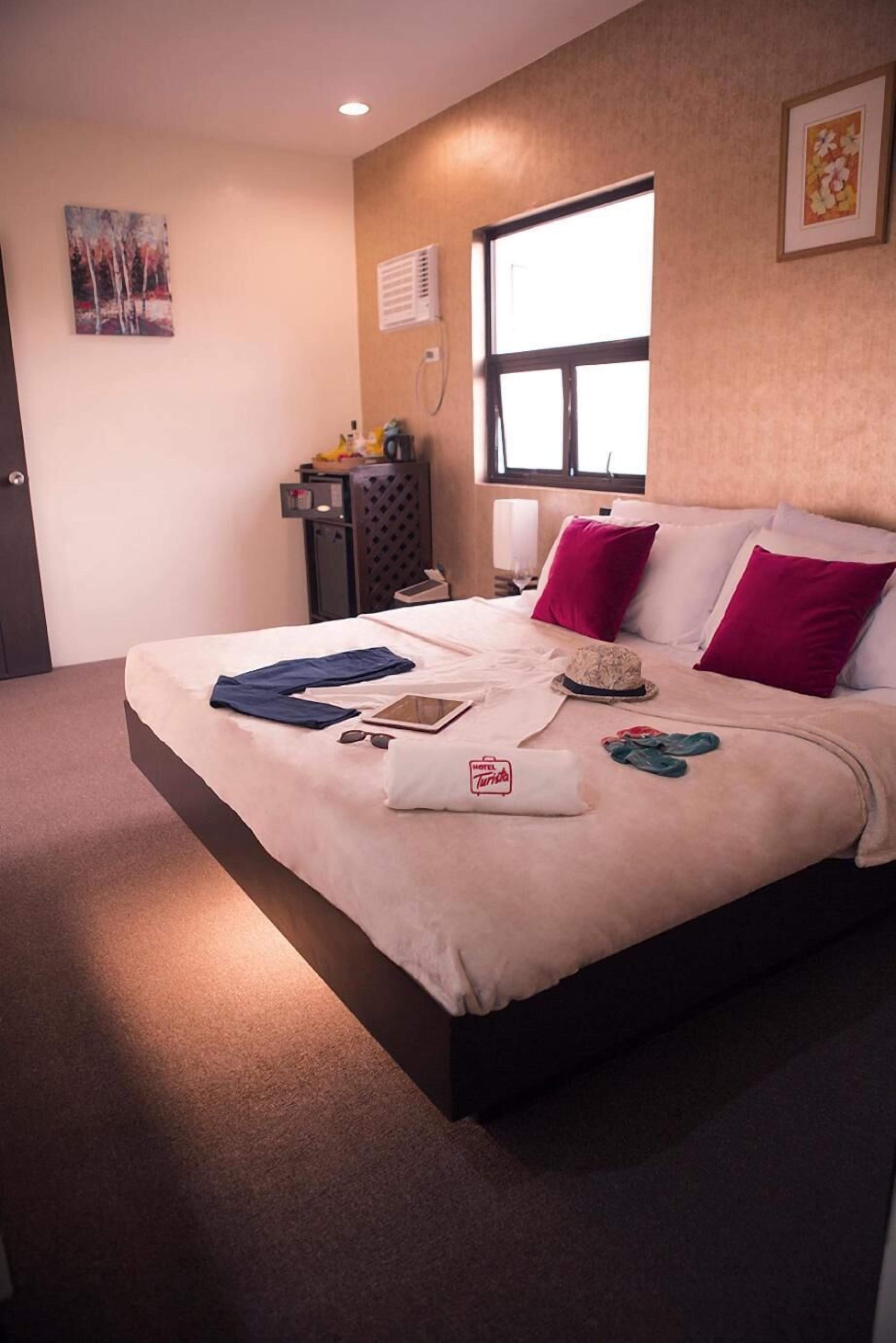 Een hotelkamer met een bed, kussens, een nachtkastje, een raam en een schilderij aan de muur.